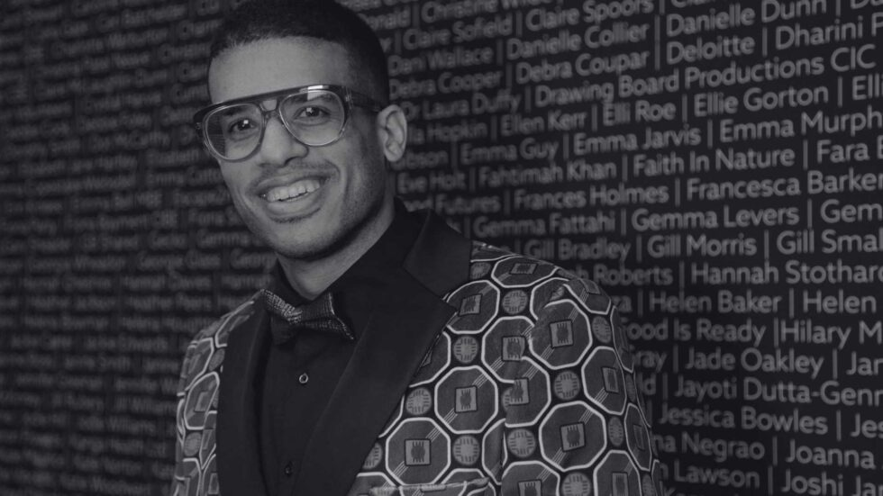 A Black man with short curly black hair, blue glasses, wearing a green jacket with a bow tie, smiling, stood against a grey background that has names written all over it