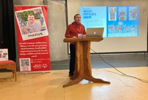 Marcus, a young man, wearing a red hoodie, standing at a lectern, giving a talk about his business, CoolArt2021 to students and staff at the Glasshouse College, Stourbridge, West Midlands. Marcus is an alumni. A slide about his business is on the screen. behind Marcus is a stand produced by Coca Cola Europacific Partners about Marcus, as part of their work with Special Olympics GB - Unified Business project.