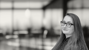 A brown haired woman with glasses in front of a blurred office.