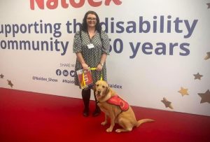 Brown haired woman standing on red carpet in front of a Naidex sign that reads ‘supporting the disability community for 50 years’. She is holding a yellow assistance dog lead with a yellow Labrador in red and yellow vest. She is wearing glasses, a black and white patterned dress, black tights and black shoes with a lanyard on.