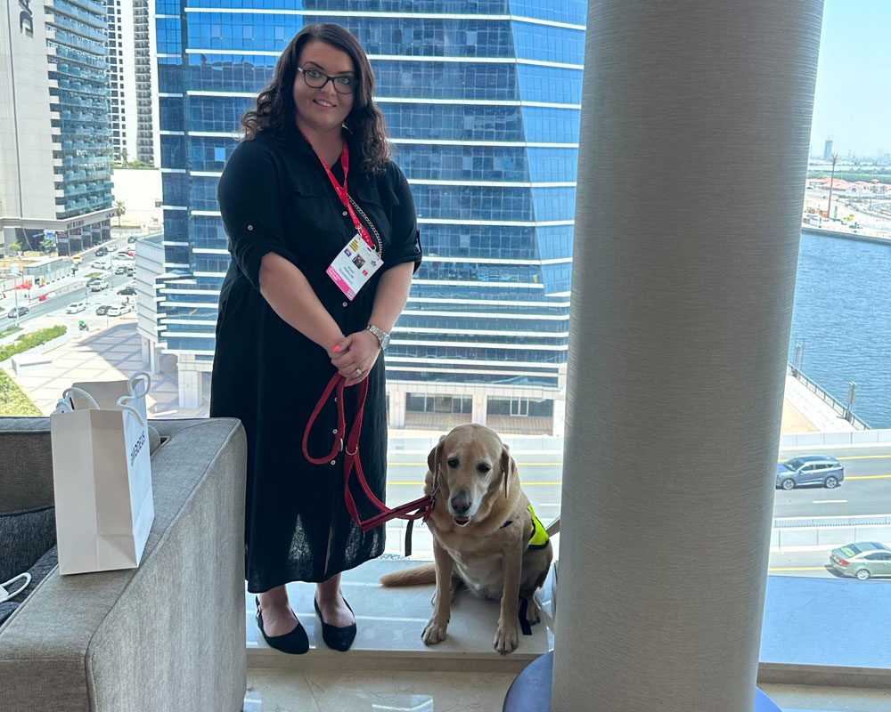 Woman with brown hair standing on a marbled floor infront of a large window with sky scraper buildings outside. To her left is an assistance dog yellow labrador wearing a yellow and red jacket arabic text. She is wearing a black dress, black shoes and glasses with red lanyard.