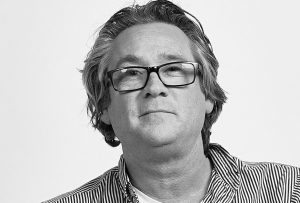 A head & shoulder portrait of a dark-haired man with glasses wearing a white t-shirt under a striped open-necked shirt against a light grey background.