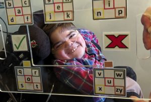 A smiling brown haired young man with nasal specks for oxygen and a plaster at the side of his face to keep them in place, wearing a blue and red checked shirt and beige chinos sat in a black wheelchair. Jonathan is facing the camera with a perspex e-tran frame with the alphabet in 5 blocks and a tick and cross stuck to it held between him and the camera. He is looking through the middle of the e-tran frame.