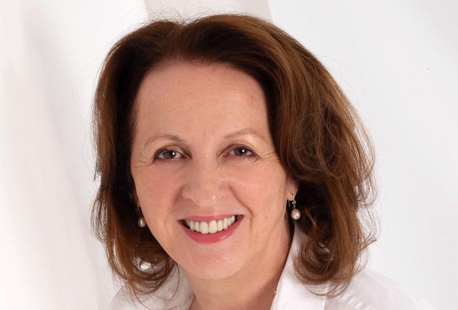 Jacqueline Winstanley, a woman with shoulder-length brown hair, is smiling warmly. She is wearing a white blouse with a collar, and pearl earrings. The background is a simple, light-colored setting, highlighting her friendly expression.