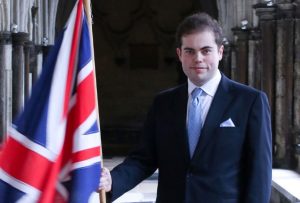 A dark haired man with blue eyes smiling, wearing a suit and holding a Union Jack flag.