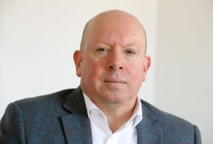 A head a shoulders picture of Julian, a white middle aged male with bald head smiling wearing a white shirt and blue jacket with a white wall behind him