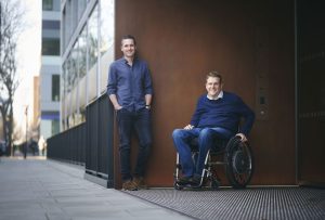 Edward Warner and James Taylor, co-founders of Motionspot, outside a building smiling. Ed is standing and James is seated in his wheelchair.