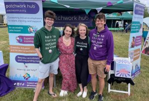 Patchwork Hub team standing smiling in front of Patchwork Hub’s stand at Parallel Windsor. Founder Beth is wearing a long red dress and her 3 colleagues are wearing Patchwork Hub tops and jumpers.