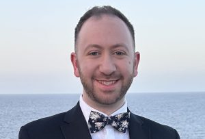 A dark haired man dressed in a black suit with blue floral bowtie, standing in front of a cruise ship balcony with the ocean in the background.