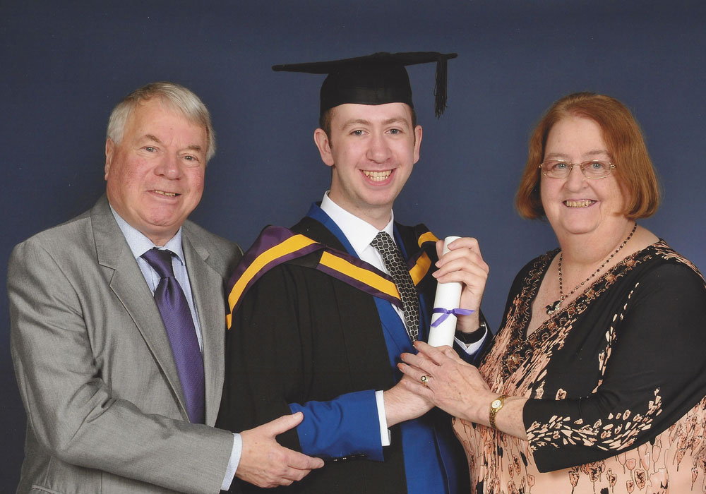 Photograph of three people at a university graduation - an older man with grey hair on the left in a suit, a younger man with dark hair in the middle in a suit wearing his university cap and gown, and an older woman with brown hair on the right wearing a blouse and trousers.