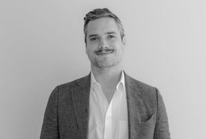 white male with a mustache and greying hair stands with his back to a white wall and wearing a white collared shirt and light blue blazer