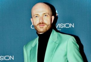 "A man with blue eyes and a shaved head, wearing a pastel green double-breasted suit jacket and black turtle beck jumper, posing for pictures on the red carpet of an event.