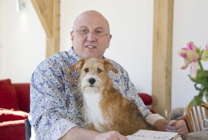 A white bald man of 60 sitting in a wheelchair, in a room with white painted walls and beams. He is wearing a blue floral shirt and has a small ginger and white dog on his knee. To the right of the image is a vase of pink flowers.