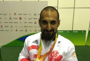 A man in a wheelchair is proudly holding a silver medal in front of a backdrop featuring various sponsor logos from the Rio 2016 Paralympic Games. He is wearing a white athletic jacket with red and blue accents, displaying the British flag and the letters "GB" for Great Britain. The medal hangs from a yellow and green lanyard around his neck, and he is smiling at the camera.