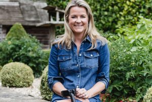 A blonde woman wearing a blue denim dress is sitting on a wall in front of a green hedge, she is looking at the camera and smiling. Leaning on her knee is her Cool Crutch.