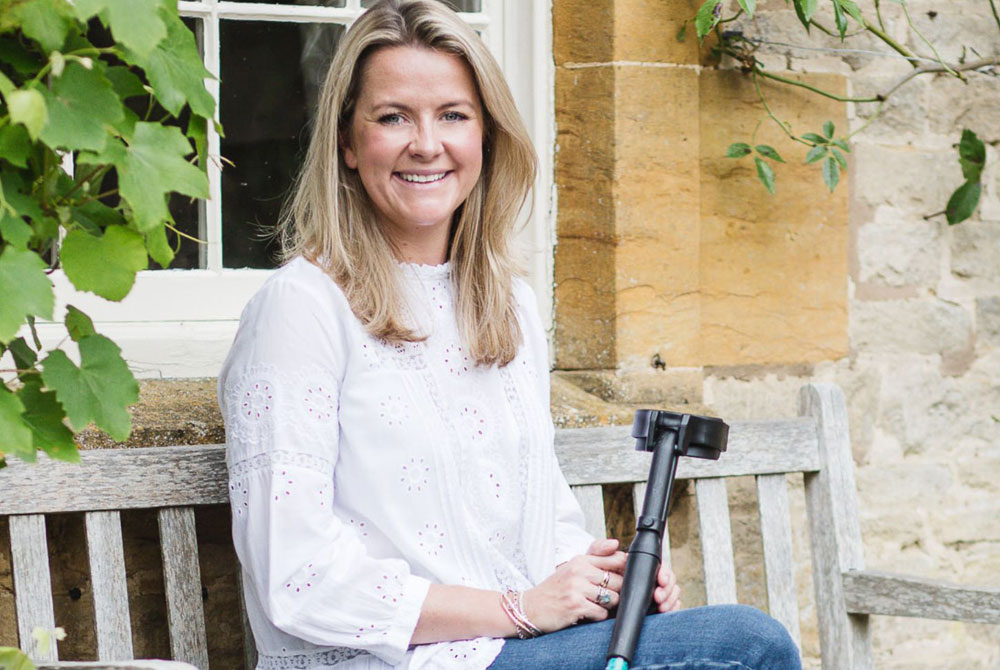 A blonde white woman sitting on a bench in front of a wall wearing a white blouse and jeans, she is looking at the camera and smiling with a single black crutch on her knee