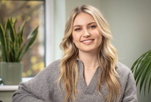 White woman with blonde wavy hair and blue eyes is sitting down smiling to camera. She is wearing a grey jumper and is sat against a green background.