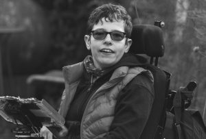 Woman with short dark hair wearing glasses sat in a power wheelchair in a garden. Mounted on the chair in front of the woman is an electronic communication device.