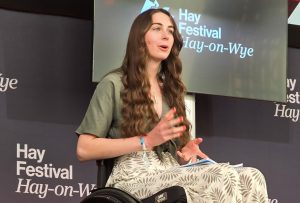 Bethany, a white woman in her twenties with long brown hair, is sat in her manual wheelchair on a stage addressing the audience.