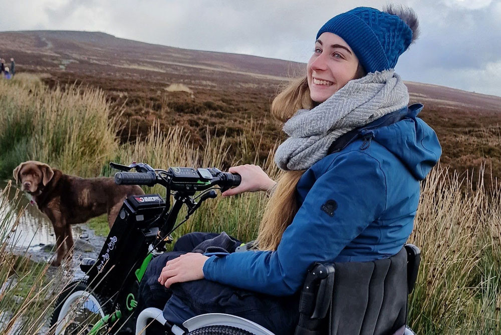 Bethany, a white wheelchair user in her 20s, is sat smiling in her wheelchair on a mountain