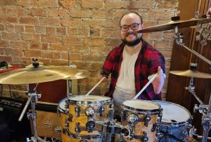 A smiling 32 year old male, short dark hair, black glasses, behind a drum kit holding sticks