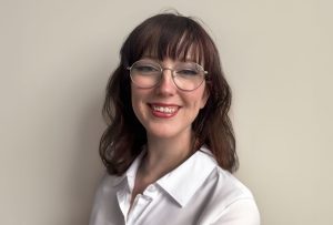 A young woman with shoulder-length brown hair and bangs is smiling warmly at the camera. She is wearing round glasses and a white button-up shirt. The background is a plain, light-colored wall.