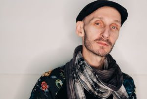 A man of white complexion, with green eyes and brown facial stubble in front of a grey wall. He has a shaved head wearing a flat cap, black jumper and a scarf around his neck.