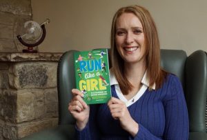 Danielle is a white woman with blonde hair and is sitting in a green armchair. She is holding her latest book Run Like A Girl up to the camera and smiling.