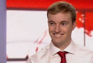 Image of a blonde man with a white shirt and red tie on, he is in a TV news studio smiling at something off camera.