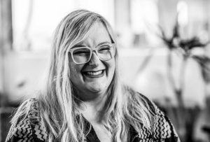 A black and white photo of Emily, a white woman wearing thick-framed glasses, laughing at the camera. Emily wears a cardigan with geometric shapes on it.