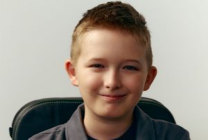 A dark blonde hair boy aged twelve sitting in his power wheelchair smiling at the camera.