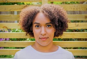 A medium brown-skinned person with a tiny afro standing in front of a garden fence.