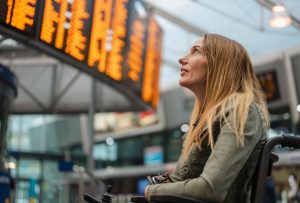 White woman with blonde hair using a wheelchair and looking up at a screen with train times on it.