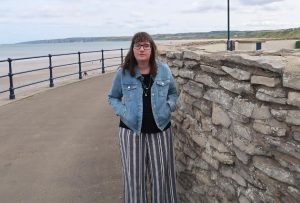 Holly stood on a promenade with the sea in the background. She's wearing a blue denim jacket and blue and white striped trousers, and has her hands in her pockets.