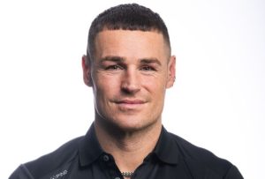 James, a dark haired man wearing a black polo top is having his photo taken professionally with a white background.