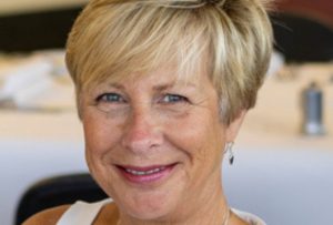 White woman with short blonde hair wearing a sleeveless cream top and smiling at the camera