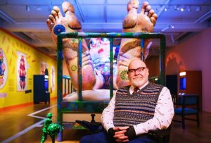 Jason Wilsher-Mills, a middle aged disabled artist, in a wheelchair, sat in front of his sculpture at the Wellcome Collection. The brightly coloured sculpture depicts a figure in a hospital bed and you can see his huge feet.