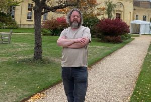 Johny Cassidy standing in a garden on a gravel path at the Radcliffe Observatory at Oxford University. He's got long curly grey hair and has a long grey beard. He's looking directly into the camera and is wearing a beige T shirt with black jeans and black boots. There's some trees and shrubs with orange flowers behind him.