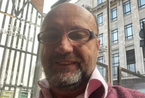 Jonathan Fletcher - Man with short hair and greying bead wearing glasses wearing pink shirt and claret jumper. In the background are two buildings one is a black and white timber framed building and a stone building.