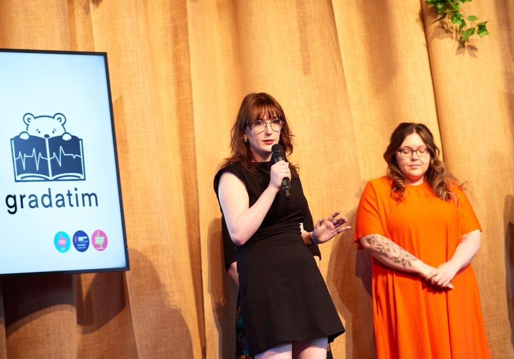 "Two women are standing on a stage in front of a beige curtain. The woman on the left is holding a microphone and speaking, wearing a black dress and glasses. The woman on the right, wearing an orange dress with a floral tattoo on her left arm, is standing with her hands clasped in front of her, looking downwards. To the left of the speaker is a screen displaying the logo for ""gradatim,"" featuring a bear peeking over a book with a heart monitor line on it, and some small icons below it.