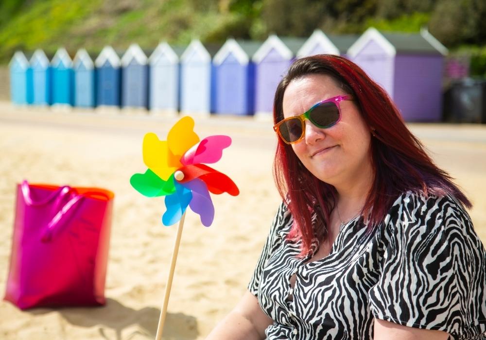 Maddy is wearing sunglasses and sat on the beach, she has red hair and is holding a plastic rainbow coloured windmill, she is wearing a zebra print dress.