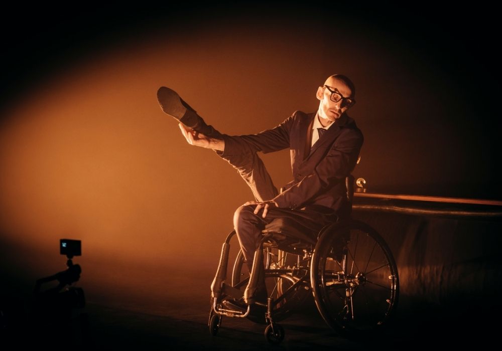 White male using a manual wheelchair in a suit and wearing glasses with a shaved head. He is lifting one leg with his arm in front of him while seated in his wheelchair.