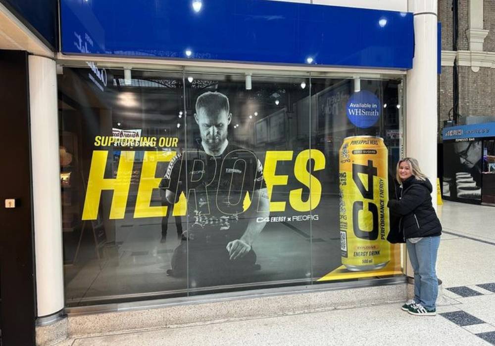 The image shows a lady with blonde hair stood outside of a WH Smith store. Covering the entire store window is an image of triple amputee Mark Ormrod in his JiuJitsu gear sat on the floor looking down after a hard training session. The image is accompanied by a picture of an energy drink from the company C4 from a campaign in partnership with the charity REORG. The image is to promote inclusivity and is intended to inspire.