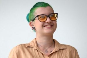 Max, a white genderqueer person with green hair, is standing against a white background and smiling. They are wearing hearing aids, glasses with orange tinted lenses, and a beige shirt.