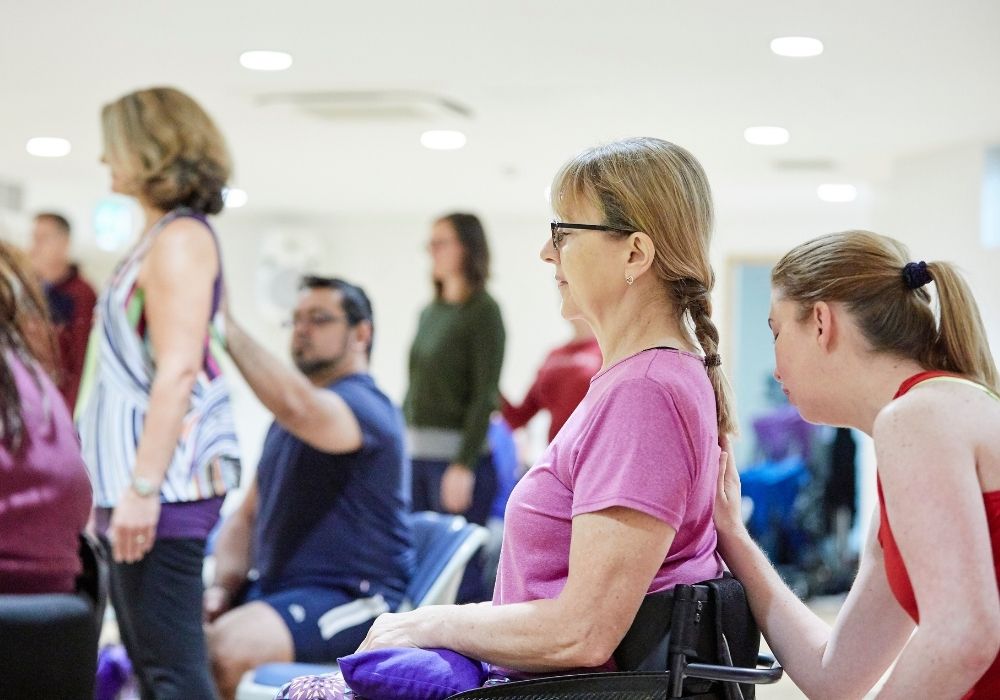 Miranda wearing a red tank top supports Siby, a wheelchair user wearing a purple t-shirt, in a seated yoga pose.