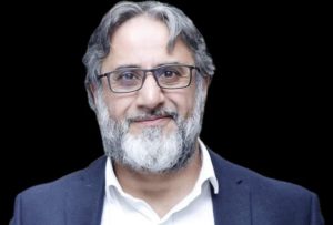 A man in glasses with a grey and white beard in a white shirt and blue jacket. He is looking at the camera and smiling.