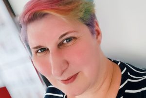 a smiling neurodivergent woman with short rainbow hair and a blue and white stripy dress leaning against a wall.