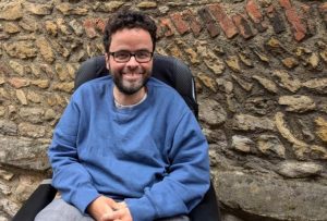 Peter is pictured seated in front of a brick wall wearing a blue top and smiling at the camera