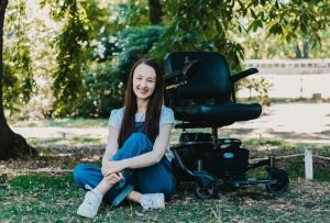 Pippa, a white woman in her twenties with long brown hair, wearing denim dungarees and sitting on the ground outdoors with one leg crossed over the other. Just behind her, you can see her black power-chair.