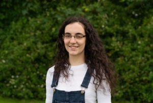 Rachel, a white woman with long brown curly hair and glasses wears a fine knit cream jumper with indigo dungarees as she smiles in front of a natural green background.